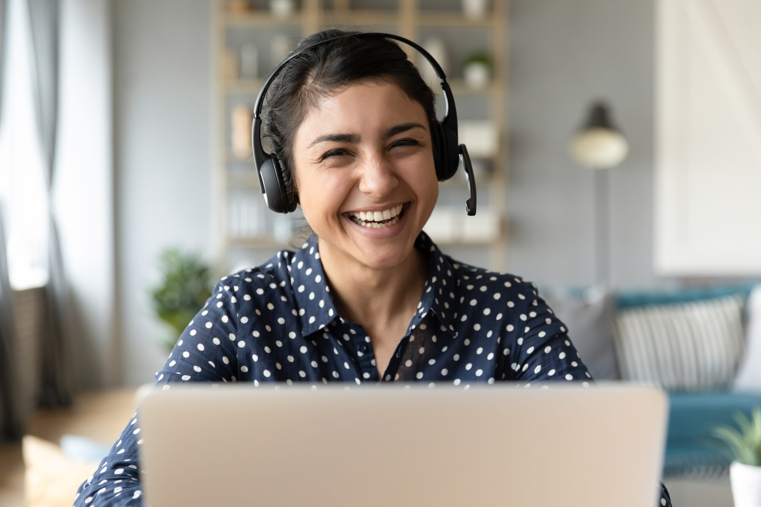 Head shot portrait smiling Indian woman wearing headphones posing for photo at workplace, happy excited female wearing headset looking at camera, sitting at desk with laptop, making video call