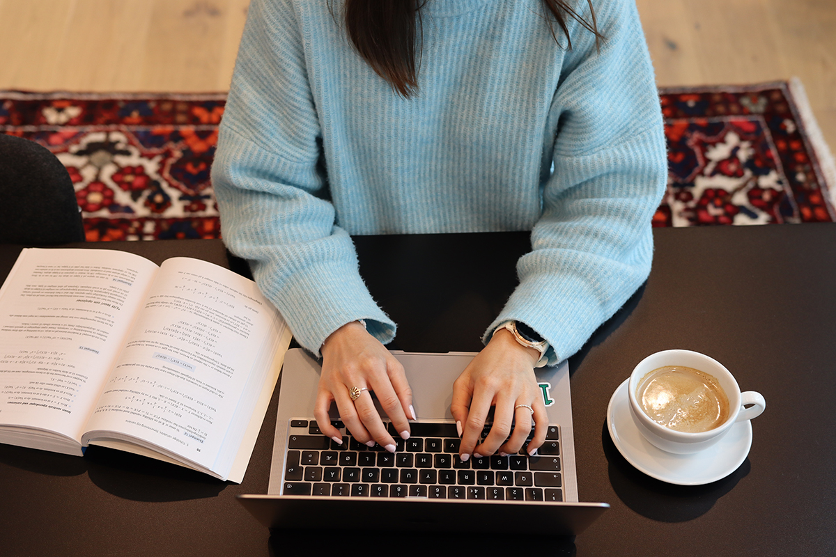 Hender på et tastatur. Pensumbok og kaffe ved siden av pc-en.